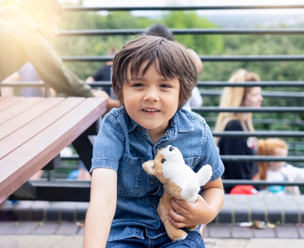 Portrait of happy little boy hugging dog toy siting on wooden bench and making funny face