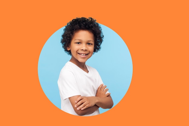 Портрет счастливого маленького мальчика в белой футболке, стоящего со скрещенными руками и улыбающегося в камеру, здорового ребенка, положительных эмоций. закрытая студия, изолированная в круглой дыре на оранжевом фоне