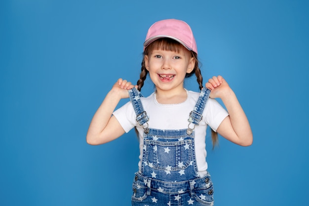 Portrait of a happy little girl 5-6 years old in a cap, on a blue wall isolate, place for text. Toothless baby. Sun protection.