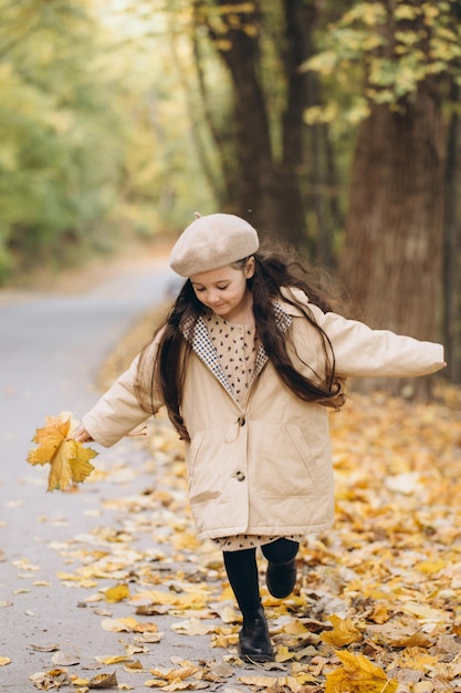 ベージュのコートとベレー帽で黄色のカエデの葉を保持し、秋の公園で過ごす幸せな少女の肖像画