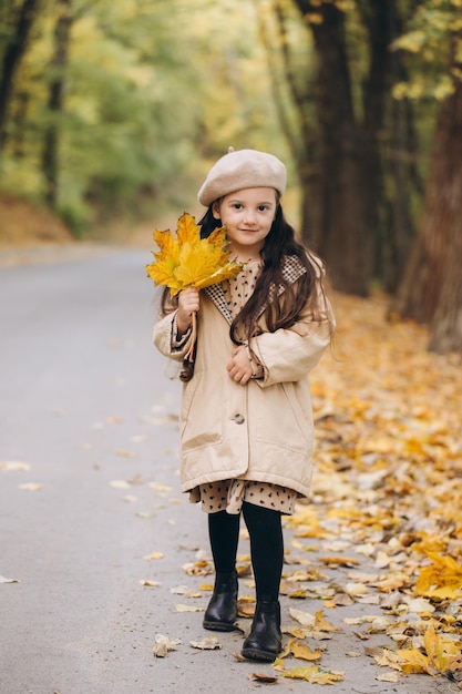 Ritratto di bambina felice in cappotto beige e berretto che tiene foglie di acero gialle e trascorre il tempo nel parco autunnale