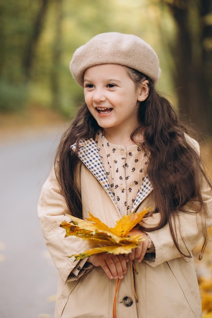 Ritratto di bambina felice in cappotto beige e berretto che tiene foglie di acero gialle e trascorre il tempo nel parco autunnale