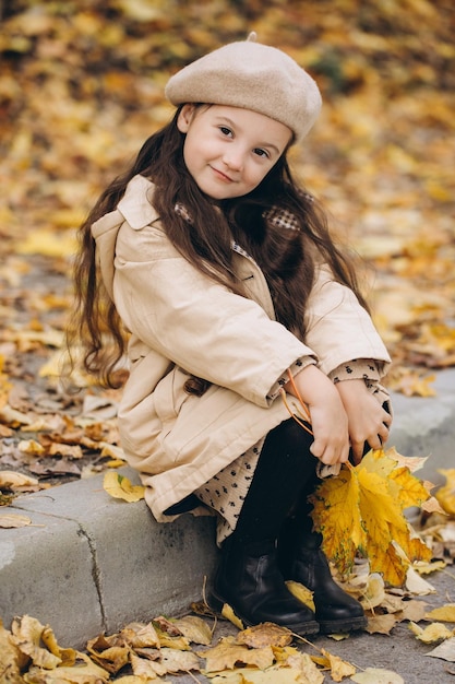 Ritratto di bambina felice in cappotto beige e berretto che tiene foglie di acero gialle e trascorre il tempo nel parco autunnale