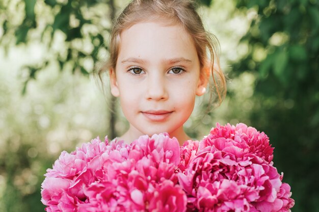 Portrait of a happy little girl, holds in hands a bouquet of pink peony flowers in full bloom