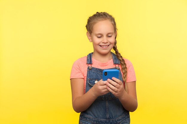 Портрет счастливой маленькой девочки, читающей смешное сообщение на мобильном телефоне, улыбающейся во время чата в социальной сети, довольной хорошим мобильным приложением в помещении студии, снятой на желтом фоне