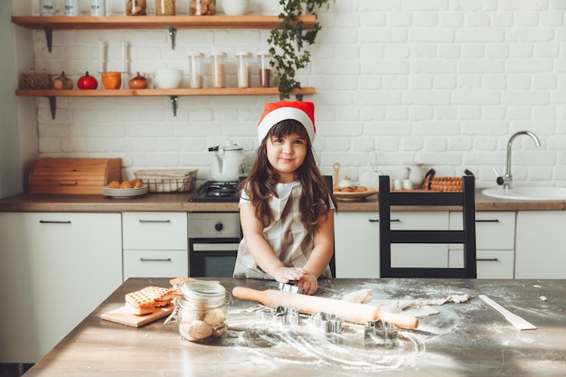 サンタ帽子をかぶった幸せな女の子のポートレート、キッチンテーブルで生地を広げ、子供がクリスマスクッキーを準備する