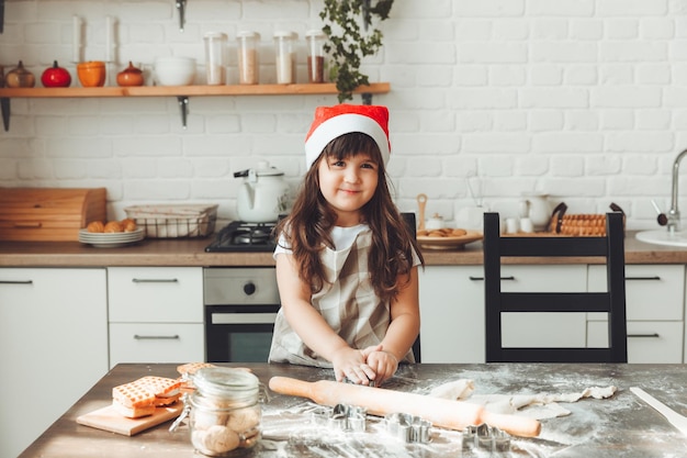 サンタ帽子をかぶった幸せな女の子のポートレート、キッチンテーブルで生地を広げ、子供がクリスマスクッキーを準備する