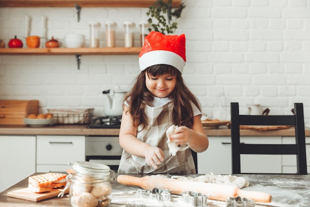 크리스마스 쿠키를 준비하는 아이가 식탁에 반죽을 굴리는 산타 모자를 쓴 행복한 어린 소녀의 초상화