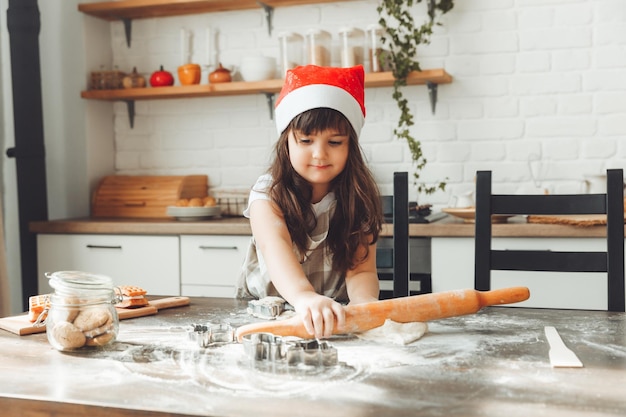 Portrait of a happy little girl in a Santa hat rolling out dough on the kitchen table a child preparing Christmas cookies