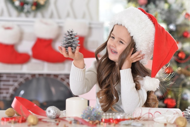 Ritratto di bambina felice in cappello di babbo natale con decorazione di pigna