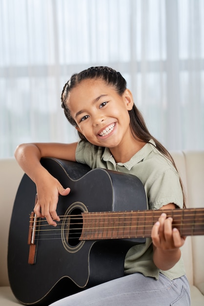 Portrait of happy little girl smiling at camera while sitting on sofa and learning to play guitar