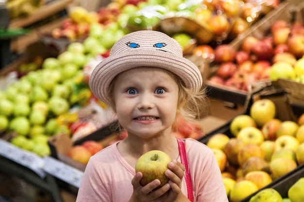 Портрет счастливой маленькой девочки с яблоком, стоящей в продуктовом магазине фруктов, глубоко думающей и смотрящей в камеру Очаровательный милый мальчик свежие фрукты