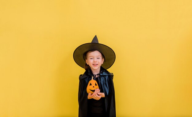 Portrait of a happy little witch in a hat with an orange pumpkin on a yellow isolated