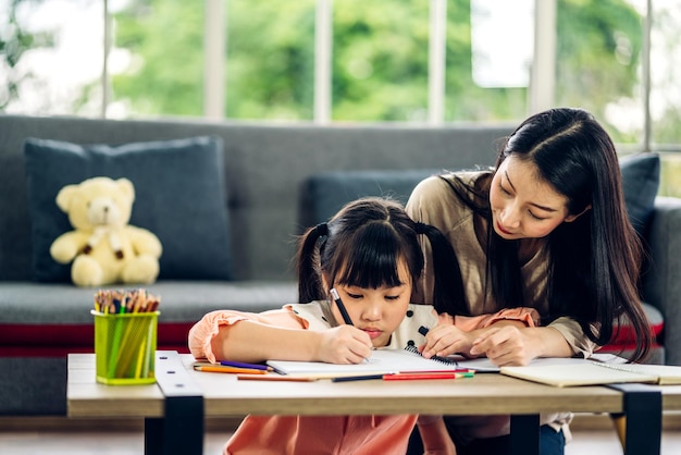 写真 肖像画幸せな愛アジアの家族の母は小さな娘に教えますアジアの女の子はテーブルで学び、勉強しますママとアジアの若い女の子は家で宿題を作る本と鉛筆で書いています教育