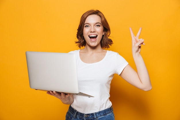 Portrait of a happy lovely girl standing isolated over yellow wall, using laptop computer