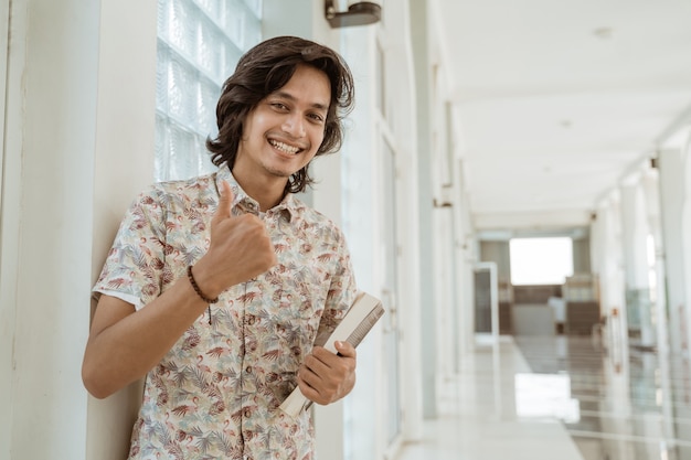 Портрет счастливого студента, держащего книгу и показывающего большие пальцы руки, смотрящего на камеру на кампусе.