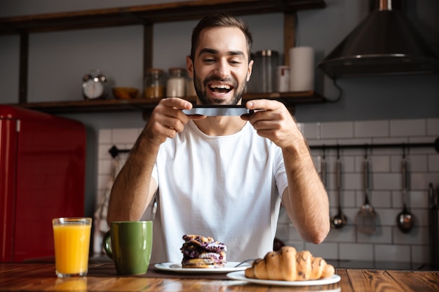Portrait of happy man 30s holding smartphone and taking photo of food while having breakfast in stylish kitchen at home