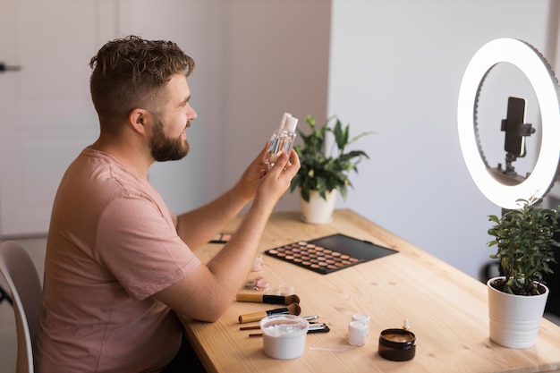 テーブルの部屋に座って、ファンデーションやカメラを見て装飾的な化粧品を勧めて話す、幸せな男の美容ブロガーのポートレート ブログと vlog のコンセプト