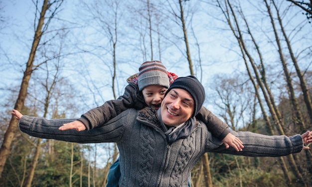 Portrait of happy man giving piggyback ride to smiling kid and raising their arms into the forest