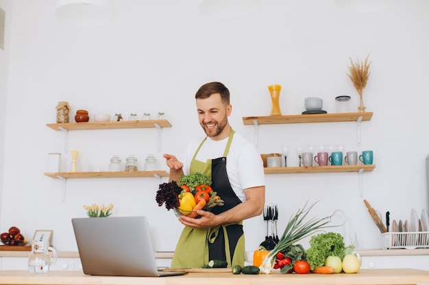 Portrait of happy man at home man cooking vegetable salad looking at camera and smiling slicing vegetables using laptop for online cooking training