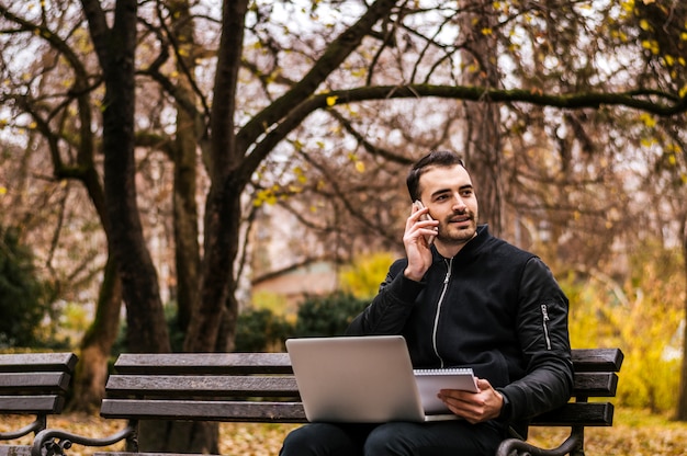 Portrait of a happy man sitting on the bench with laptop computer and talking on the phone outdoors