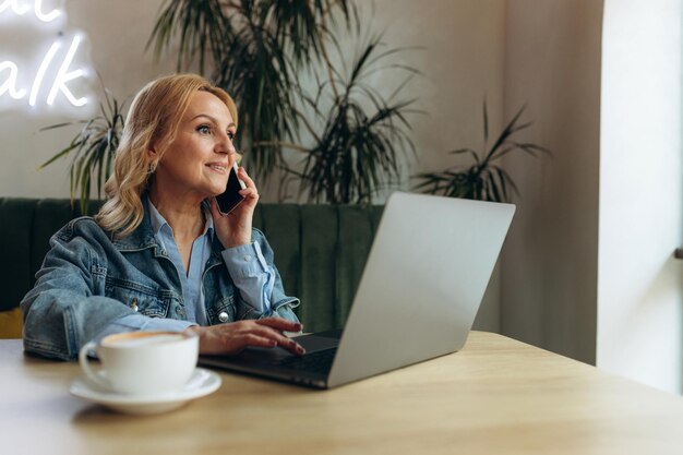 Портрет счастливой зрелой женщины, сидящей в кафе, работающей на ноутбуке и разговаривающей по мобильному телефону, пожилой женщины в классической одежде Концепция фрилансера