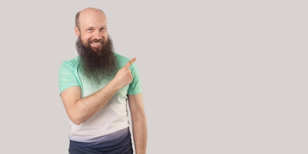 Portrait of happy middle aged bald man with long beard in light green tshirt standing looking with toothy smile and pointing at wall empty copyspace indoor studio shot isolated on grey background