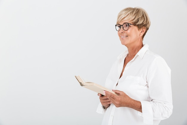 Portrait of happy middle-aged woman wearing eyeglasses holding book and looking aside isolated over white wall in studio