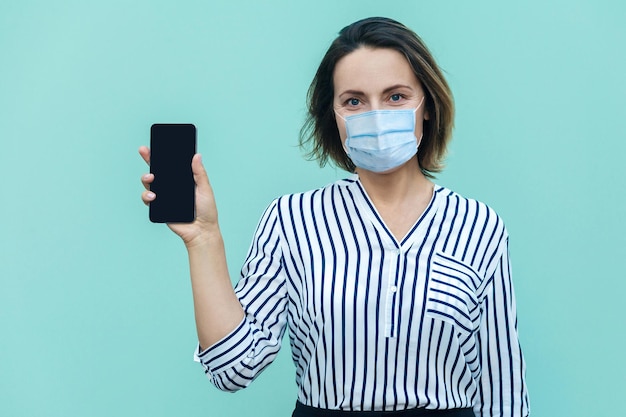 Portrait of happy middle aged woman with surgical medical mask standing holding and showing smart