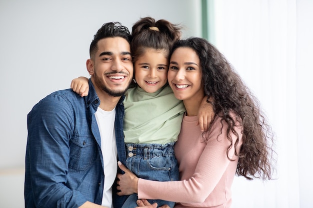 Ritratto di felice famiglia mediorientale di tre in posa insieme a casa, amorevole giovani genitori arabi e la loro piccola figlia carina abbracciando vicino alla finestra e sorridendo alla telecamera, primo piano, spazio di copia