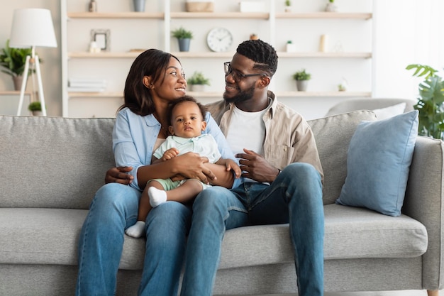 Ritratto di felice coppia afroamericana millenaria con figlio piccolo seduto sul divano abbracciandosi guardandosi e sorridendo, genitori felicissimi che posano abbracciando un bambino carino