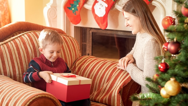 Portrait of happy mother giving Christmas gift i beautiful box to her little son sitting in armchair next to beautiful Christmas tree. Perfect image for winter holidays and celebrations