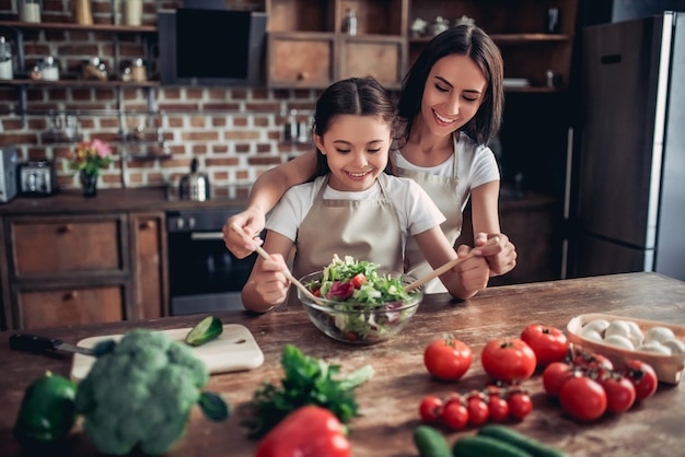 Portrait of happy mother helping her daughter tossing the salad in the bowl on the kitchen