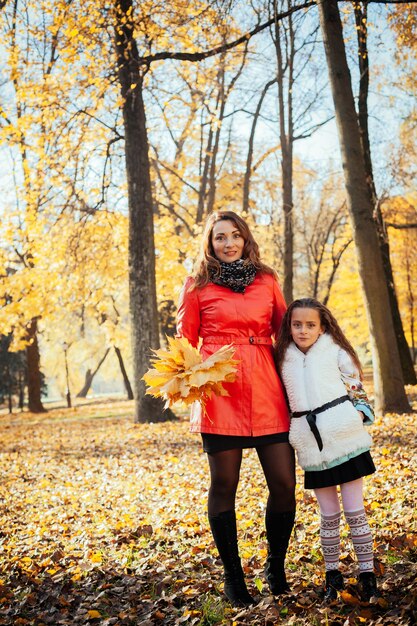 Portrait of happy mother and her daughter in autumn forest at sunset with tree leaves in her hands