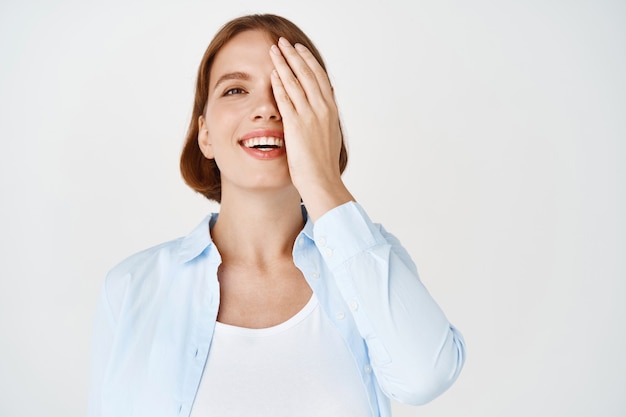 Portrait of happy natural woman cover half of face and smile , standing in blouse against white wall