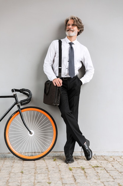 Portrait of happy old businessman in eyeglasses looking aside while standing with bicycle over gray wall outdoors
