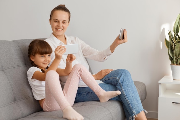 Portrait of happy optimistic woman wearing white shirt and jeans sitting on sofa with her daughter and looking at child's phone screen while having video call or broadcasting livestream.