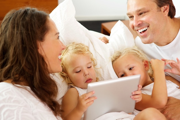 Portrait of happy parents with little daughters lying on bed using digital tablet