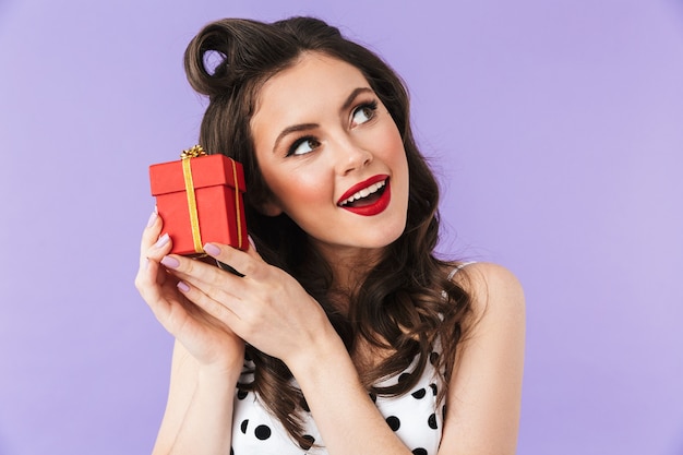 Portrait of happy pin-up woman in vintage polka dot dress smiling while holding red present box isolated over violet wall