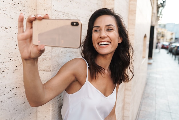 Photo portrait of happy pleased woman smiling and taking selfie photo on smartphone while standing over wall on city street