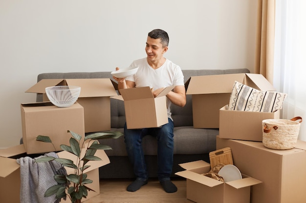 Portrait of happy positive man wearing white casual T-shirt and jeans, sitting on sofa, unpacking boxes after moving, holding box and taking out plate with optimistic facial expression.