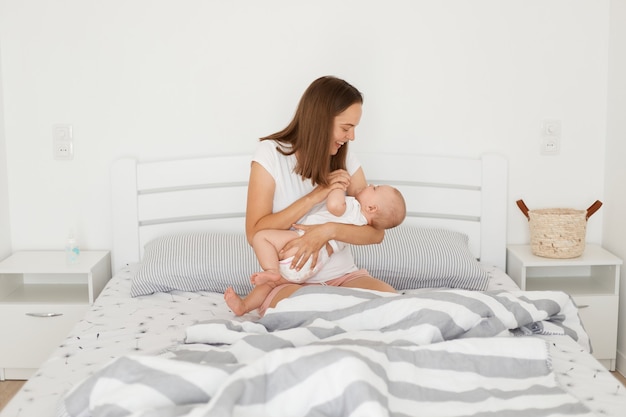 Ritratto di felice giovane madre ottimista positiva che tiene in mano un bambino piccolo, tocca il braccio minuscolo del bambino, gioca con il suo affascinante bambino carino, maternità felice.