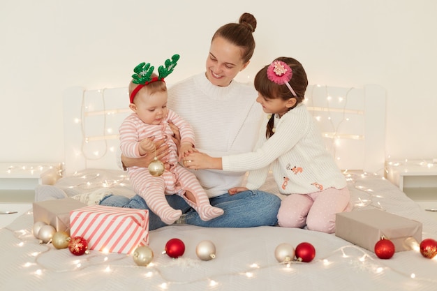 Portrait of happy positive woman posing with her children, family wearing casual attires, sitting on bed decorated with present boxes and baubles, enjoying winter holidays.