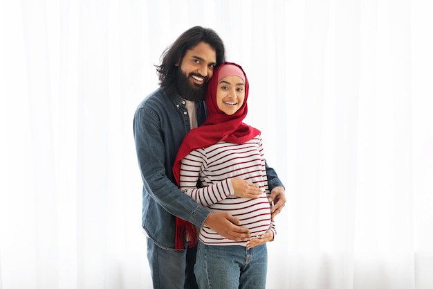 행복 한 임신 한 무슬림 부부 의 초상화 가 집 의 창문 근처 에 포즈 를 취 하고 있다