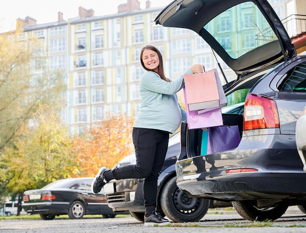 Portrait of happy pregnant woman standing near open trunk of car at parking lot in yard of apartment buildings Gravid lady holding several bright paper bags bending her leg at knee