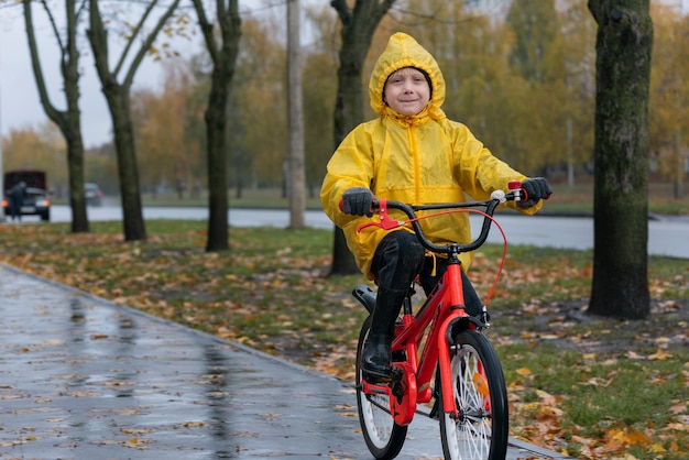 黄色のレインコートで幸せな未就学児の肖像画。少年は街中の雨の中、秋の公園で自転車に乗る。