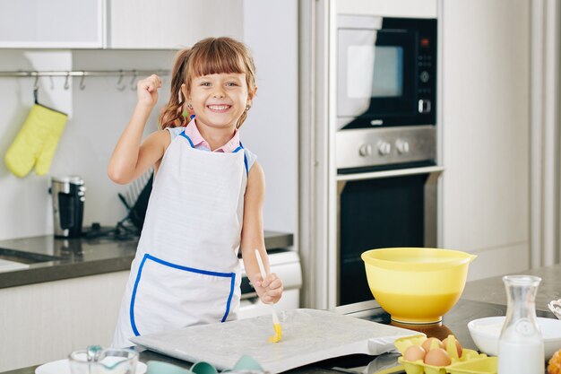 Portrait of happy preteen girl covering baking sheet with soft butter before putting dough on it