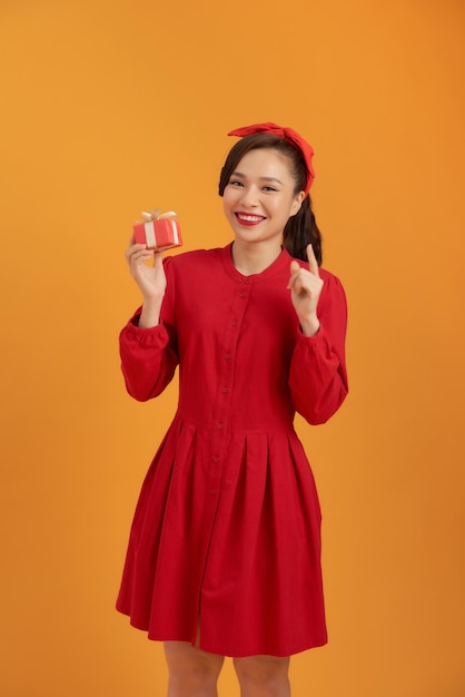 Portrait of a happy pretty asian woman holding present box while standing and looking at camera isolated over orange background