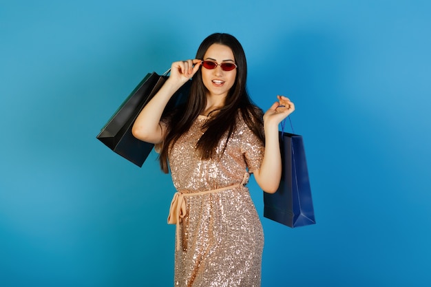 ショッピングバッグを保持し、青い背景で隔離のカメラを見ているエレガントなドレスと赤いサングラスで幸せなかわいい女の子の肖像画。
