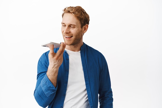 Portrait of happy redhead guy record voice message and smiling holding smartphone near mouth chatting translate his words in mobile application standing over white background